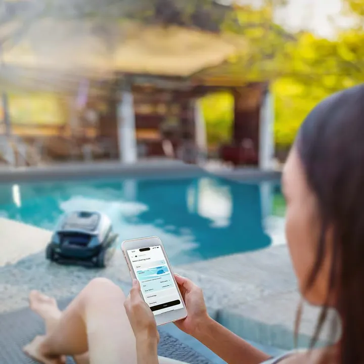 A woman by the pool checks the Dolphin app while an EON robot cleans